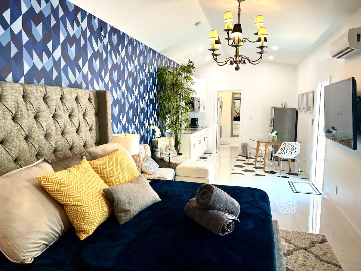 A cozy studio space features a plush bed adorned with decorative pillows, complemented by a patterned accent wall in shades of blue. A stylish chandelier provides overhead lighting, while a seating area and modern entertainment unit enhance the living space. Large windows invite natural light into the room.