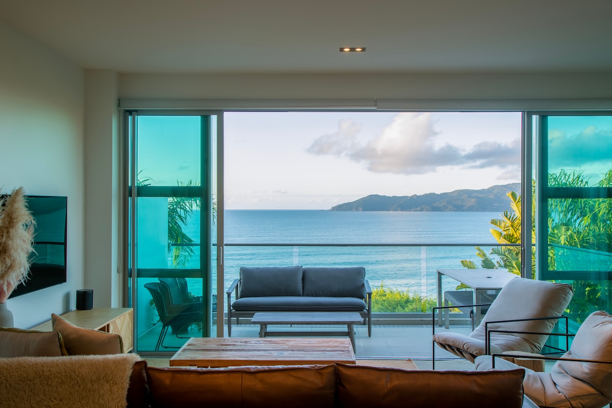 The spacious living area features large glass doors that open to stunning sea views. The decor includes a comfortable sofa and armchairs, with a wooden coffee table at the center. Natural light illuminates the room, enhancing the serene coastal ambiance.