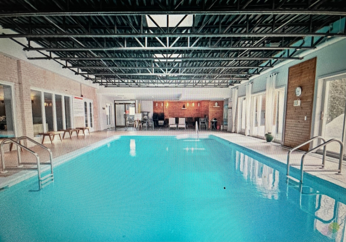 An indoor swimming pool is featured, surrounded by large windows allowing natural light. Comfortable seating areas are visible in the background, with a wooden accent wall adding warmth. The ceiling is high and supported by a metal structure, enhancing the open atmosphere.