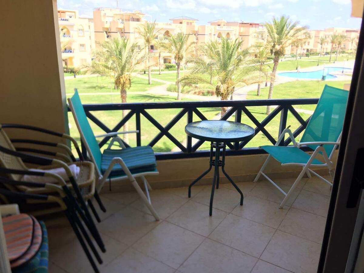 A balcony seating area is presented with a round table and a mix of lounge chairs. A view of palm trees and a green lawn is visible beyond the railing, with a swimming pool situated nearby. Natural light brightens the tiled floor.