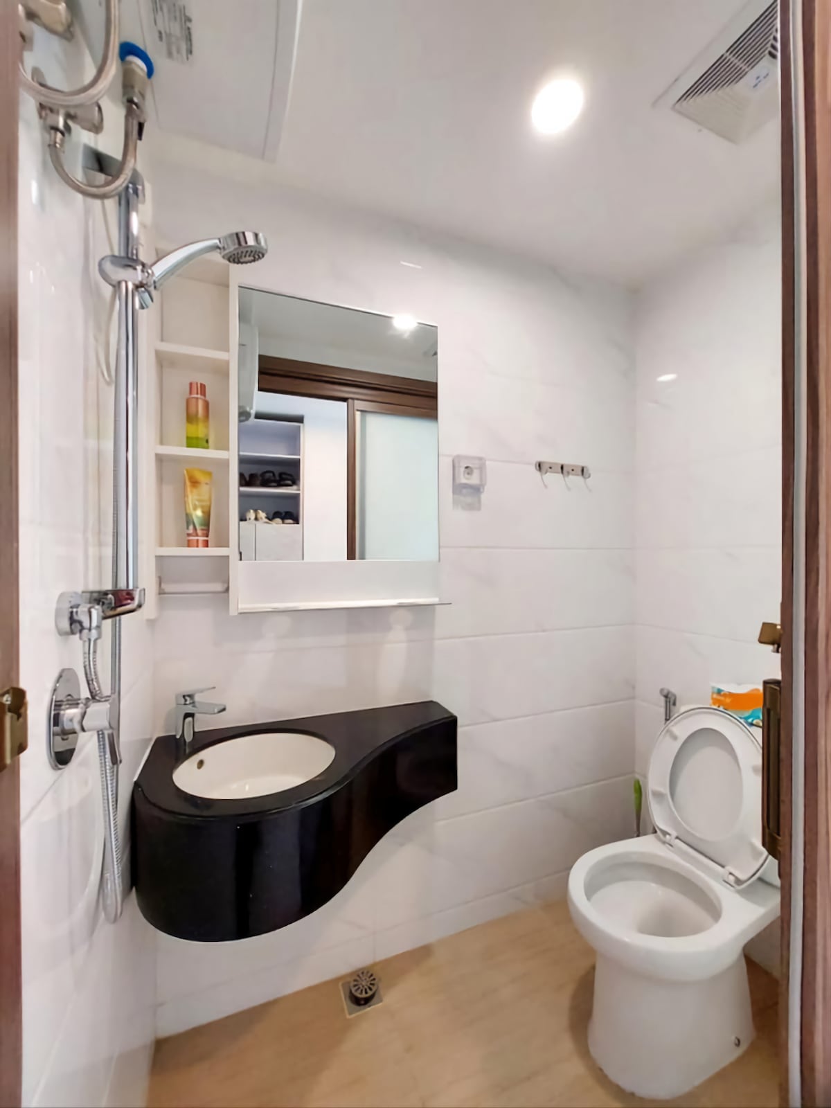 A modern bathroom features sleek white tiles, a wall-mounted mirror above a curved black sink, and a compact toilet. Shelves are integrated for toiletries, and warm lighting enhances the space. A clear separation between the wet and dry areas is evident.