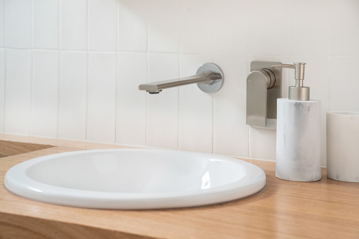 A clean and modern bathroom sink area features a round white basin set within natural wooden countertops. A sleek chrome faucet and a minimalist soap dispenser are positioned nearby against a backdrop of textured white tiles.