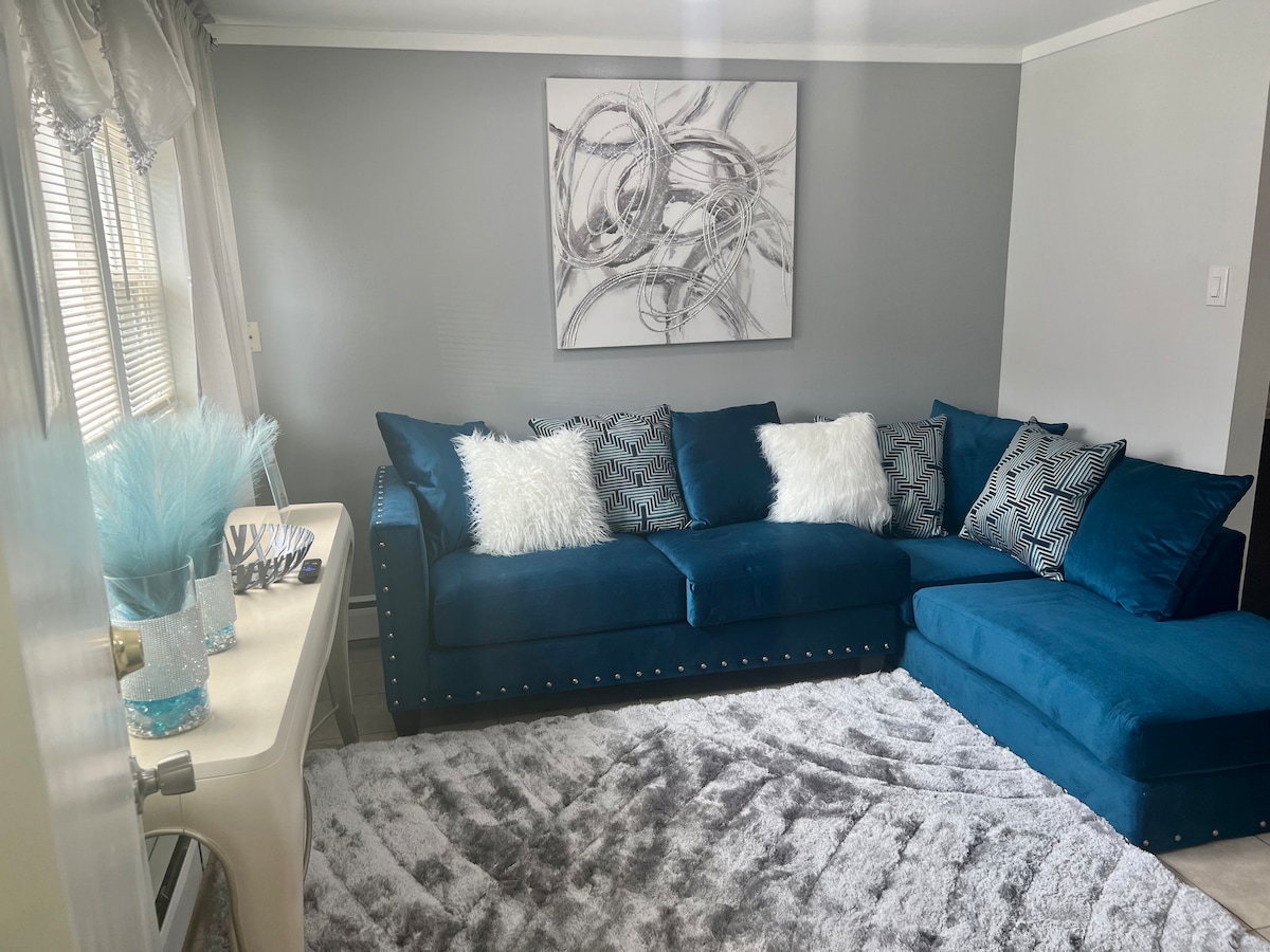 A stylish seating area features a spacious blue sectional sofa adorned with decorative pillows in varying patterns. A soft, light grey area rug lies beneath, complementing the blue tones. A modern piece of wall art is displayed above, contributing to the apartment's contemporary design.