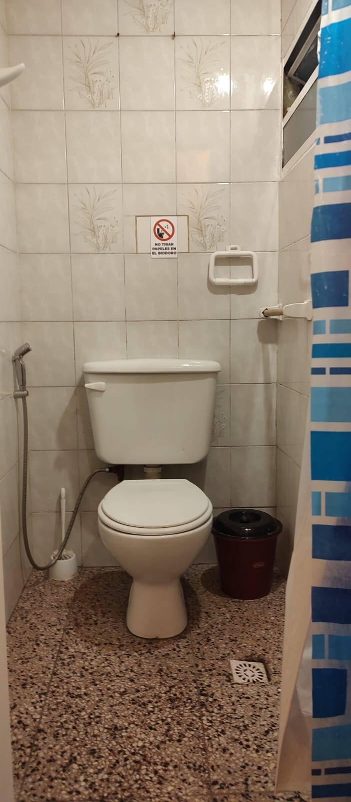 A compact bathroom is featured, showcasing white tiled walls and a clean toilet. A wastebasket sits on the floor, while a shower curtain with a blue pattern adds a pop of color. An informational signage is mounted on the wall, ensuring guest comfort.