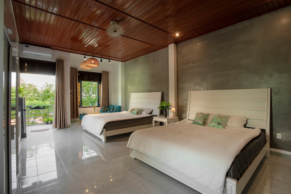 A spacious bedroom features two large beds complemented by light linens and decorative pillows. Large windows provide natural light and views of the greenery outside. A cozy seating area is visible near the window, enhancing the room's relaxing ambiance.