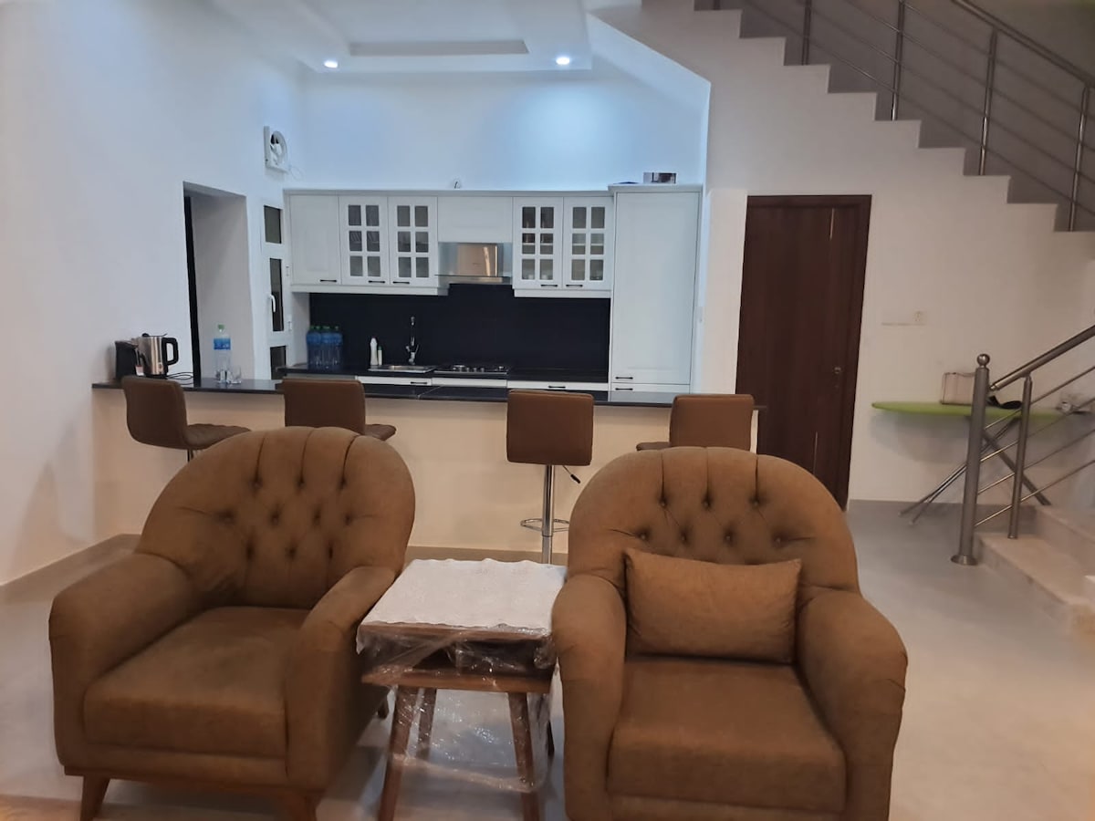 The open living area features two plush armchairs with a neutral upholstery surrounding a small wooden table. A well-equipped kitchen is visible in the background, displaying light cabinetry and a dark countertop. A staircase leads to the upper level, adding depth to the space.