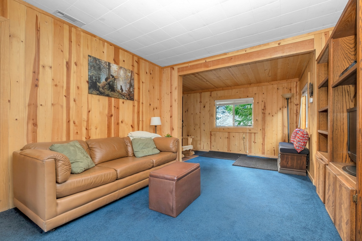 A spacious living area is enhanced by wooden paneling and plush blue carpeting. A tan leather sofa with green cushions is positioned beside a small ottoman. Natural light enters through a nearby window, and a cozy reading nook is visible in the corner.