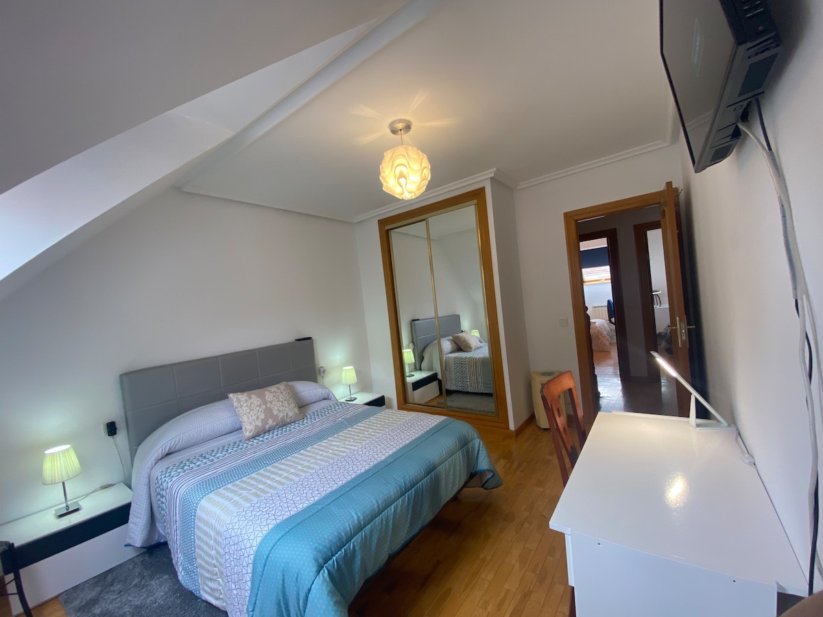 A well-lit bedroom features a spacious bed with a light blue blanket, accompanied by bedside tables with soft lamps. A full-length mirror reflects the room's layout, which includes a desk and a flat-screen television mounted on the wall. Natural light is invited through the window.