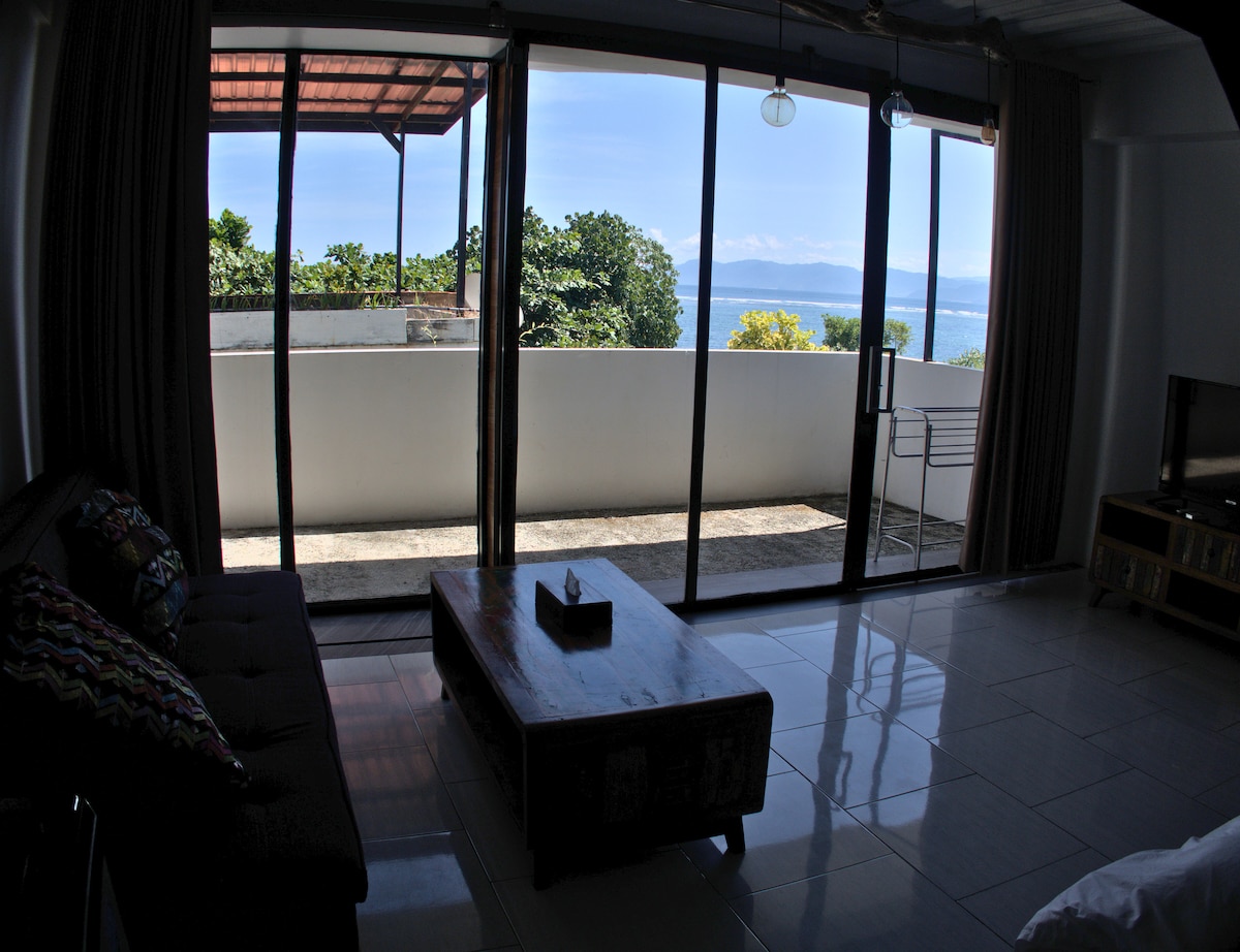 A spacious living area features large glass doors that open to a balcony with panoramic views of the landscape and water. Natural light illuminates the room, highlighting a comfortable sofa and a wooden coffee table. The décor blends functionality with a touch of style.