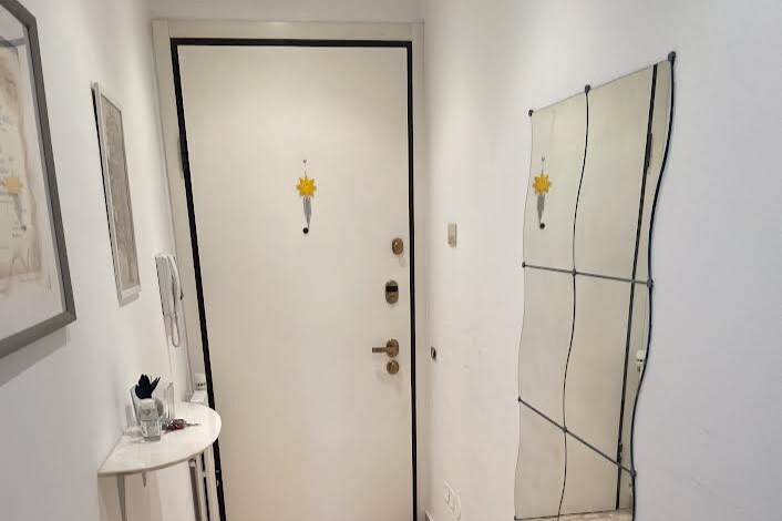 The entrance features a white door with an ornamental touch, complemented by a slender, wavy mirror. A small table holds decorative items, and a phone is mounted on the wall. Soft lighting contributes to the welcoming ambiance of the space.