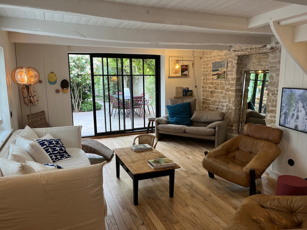 A well-lit living area features a mix of seating options, including a two-seater sofa and a leather chair. A wooden coffee table is centrally positioned, while large glass doors open to the garden, allowing natural light to brighten the space. Textured walls and wooden flooring add warmth.