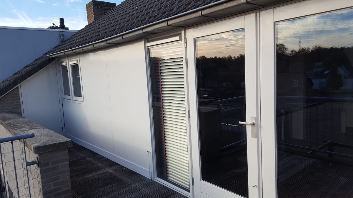 A spacious balcony is featured, surrounded by a low railing. Large glass doors provide access to the interior, reflecting the surrounding area. Windows are equipped with blinds, allowing for light control. The roofline and overhang are visible, creating a shaded area on the balcony.
