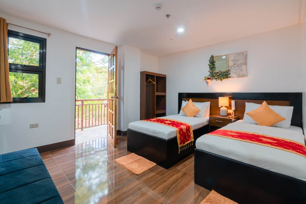 A spacious room features two single beds adorned with orange accent pillows and a red throw. Natural light filters through the large window, which offers access to a wooden balcony. A wooden wardrobe and a soft seating area complete the inviting space.