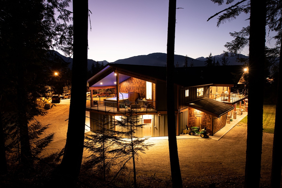 The exterior of the Crowbar Lodge is illuminated against the evening sky, highlighting the spacious structure with large windows. The surrounding trees frame the building, and lights from the parking area add warmth to the inviting setting.