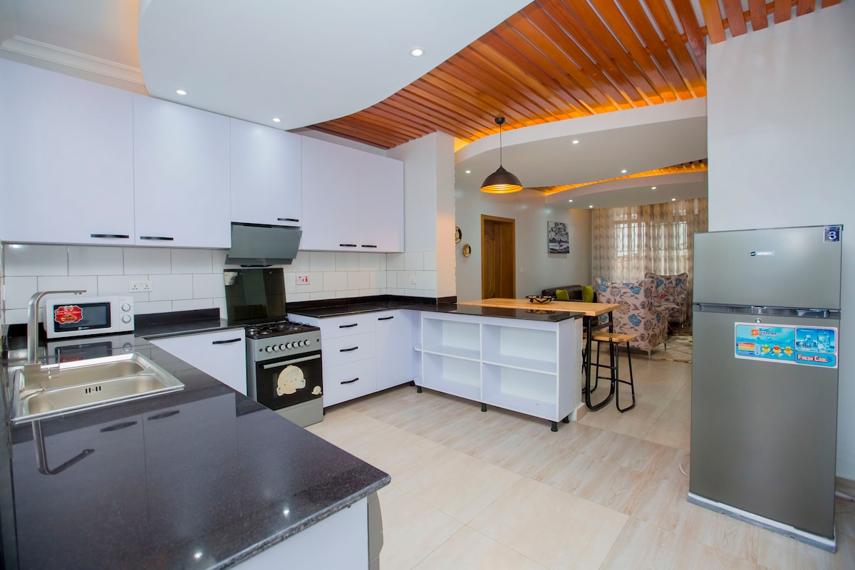 The kitchen features modern cabinetry with sleek finishes, complemented by a black countertop. An island with bar stools provides additional seating, while a refrigerator and stove complete the space. Natural light illuminates the area, enhancing the open layout.