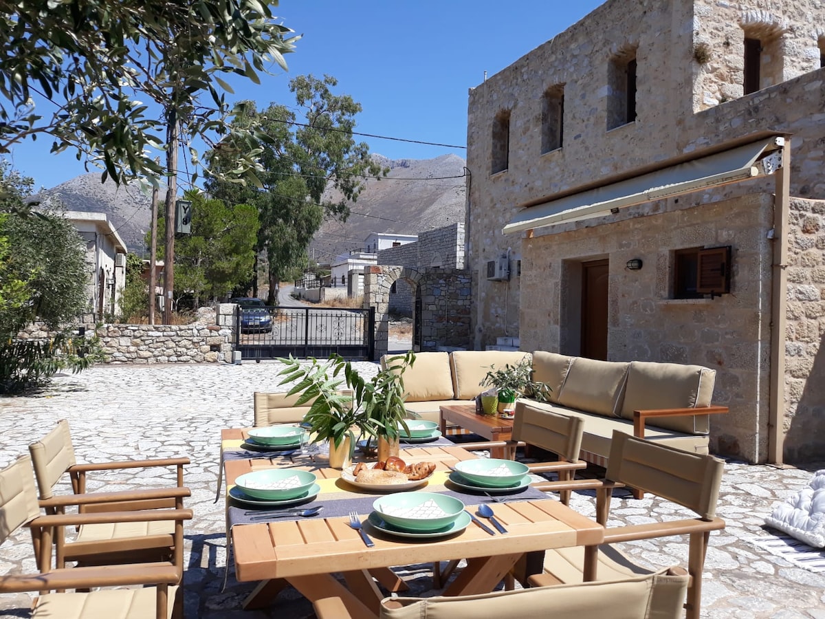 An outdoor dining area is arranged around a wooden table adorned with green plates and a centerpiece of greenery. Comfortable seating is provided by beige lounge chairs. Stone buildings and trees create a serene backdrop, with a clear view of gently sloping mountains in the distance.