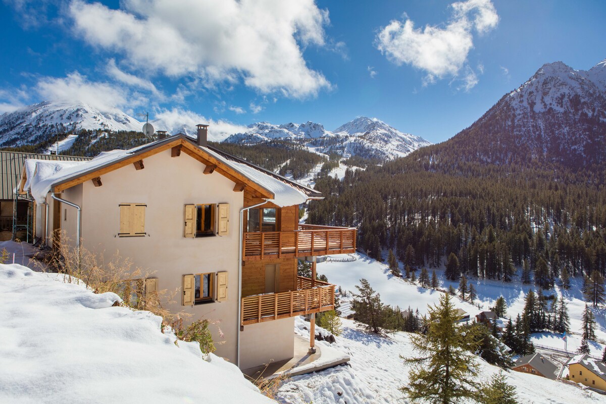 The chalet is set against a backdrop of snow-covered mountains, showcasing a blend of wooden architecture and large balconies. Sunlight reflects off the snow, enhancing the serene winter landscape. Pines dot the foreground, adding to the natural beauty surrounding the property.