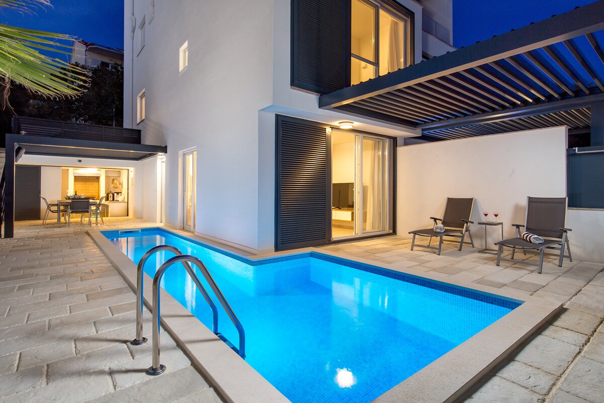 An illuminated patio features a private heated pool surrounded by light-colored tiles. Two lounge chairs are positioned near the water, while an outdoor dining area is visible in the background. Large windows provide views into the home, enhancing the inviting atmosphere.