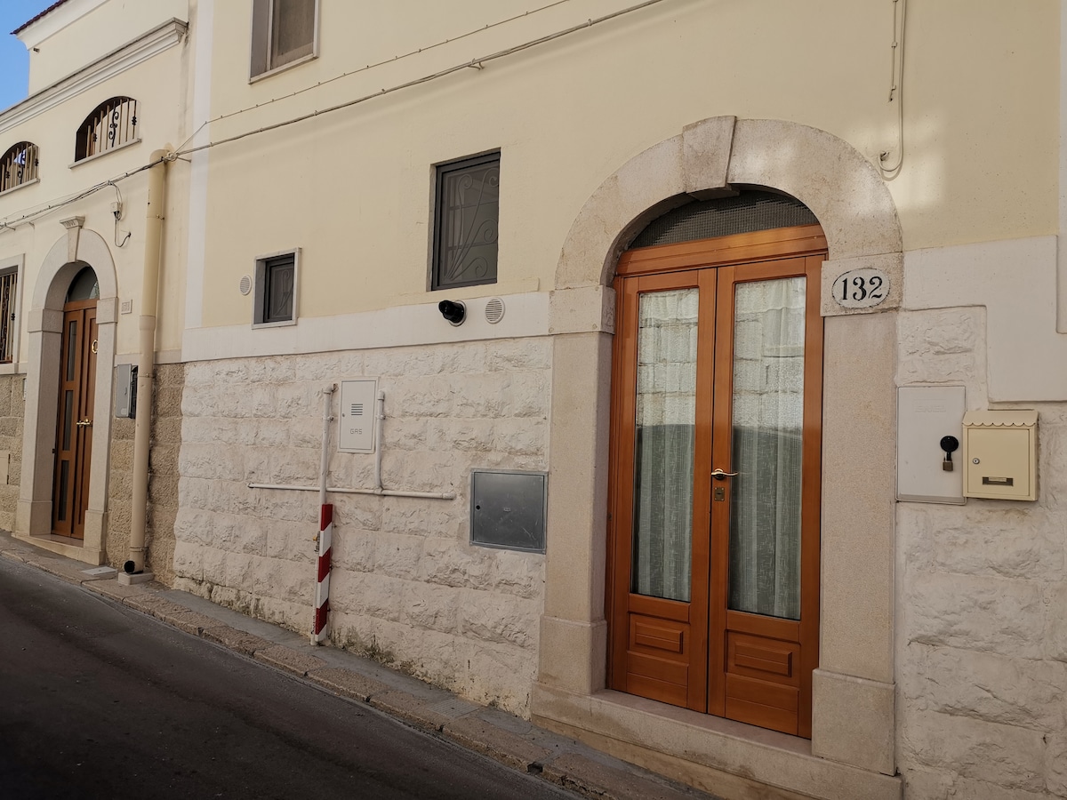 The exterior view features a stone façade with a pair of wooden front doors, each framed by an arched entrance. The street setting includes a nearby wall mailbox and a numbered plaque indicating street address 132, with simple window designs above the doors.