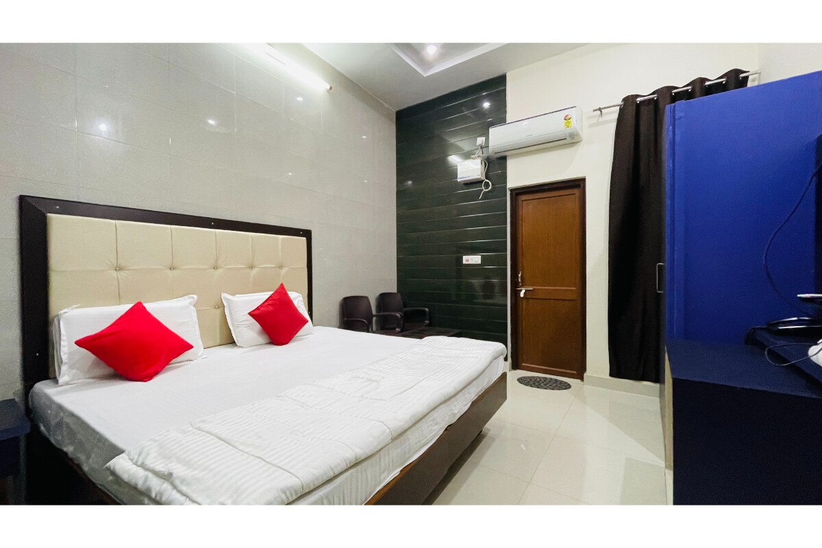 A spacious room features a large bed with white linens and red accent pillows. Neutral-toned walls and polished tile flooring enhance the clean aesthetic. A wooden door leads outside, while a small seating area is visible on one side. An air conditioning unit and a TV are included.