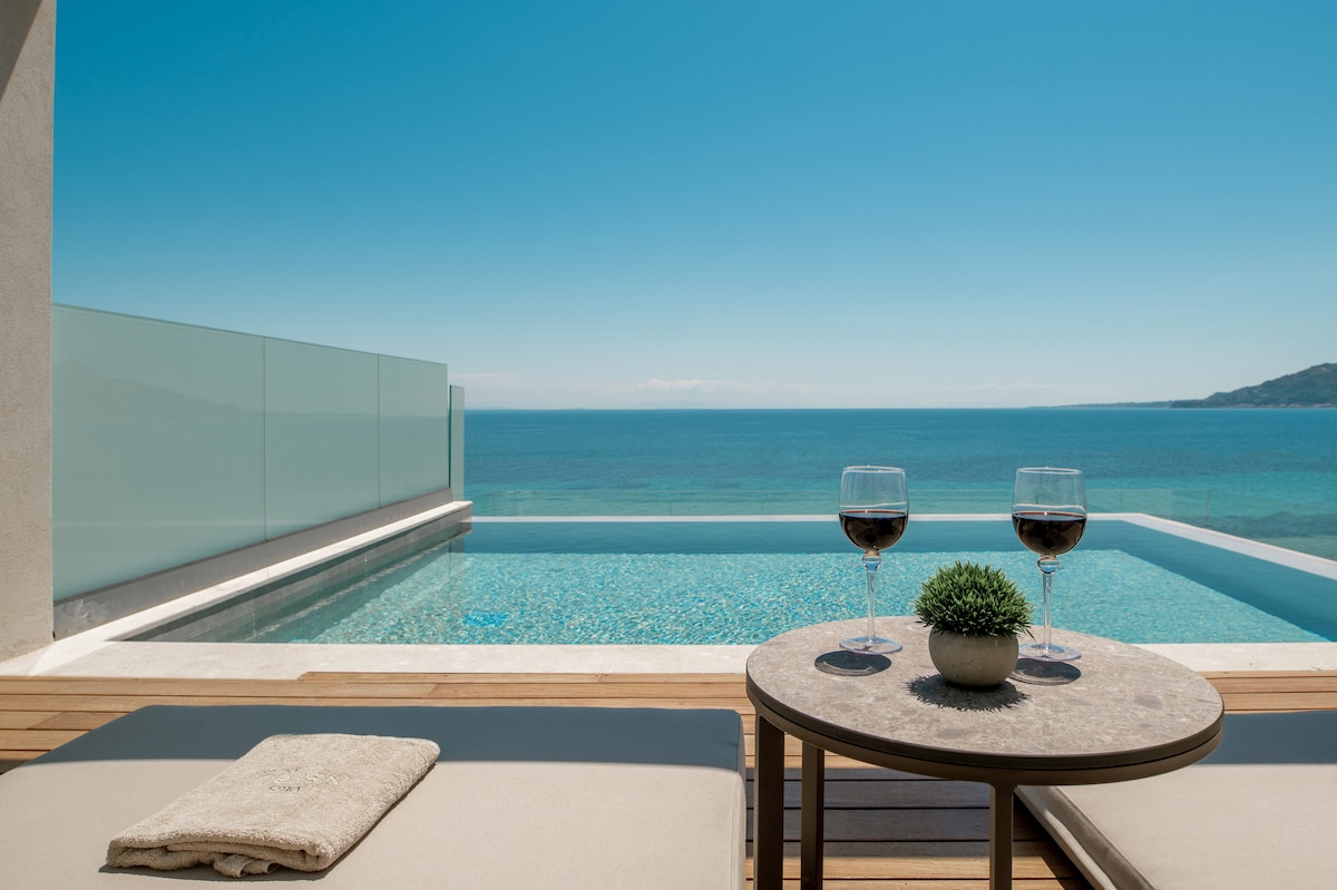 A private infinity pool extends over the edge, offering unobstructed views of the serene Ionian Sea. Two glasses of red wine rest on a stone table, accompanied by a small potted plant, while sun loungers invite relaxation under clear blue skies.