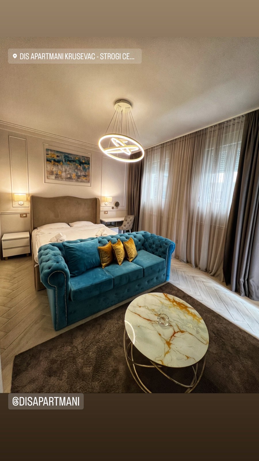 A spacious sleeping area features a large, upholstered bed complemented by a stylish blue sofa adorned with decorative pillows. A round marble coffee table with a subtle golden accent sits in the center, while sheer curtains allow natural light to filter in.