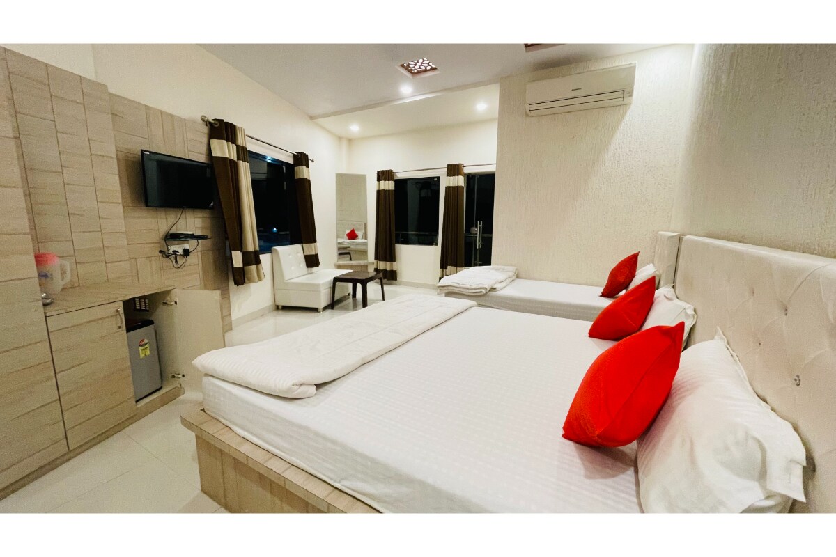 A spacious hotel room features two beds adorned with red accent pillows. Natural light enters through large windows, which are dressed with coordinating curtains. A small sitting area includes a white chair and table. A television is mounted on the wall alongside a compact refrigerator.