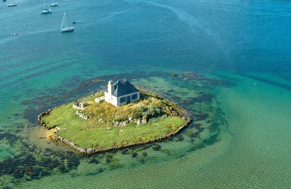 An isolated house is positioned on a small island surrounded by calm waters. The vibrant turquoise sea contrasts with patches of rocky shore and lush greenery. Sailboats can be seen in the background, enhancing the tranquil coastal scene.