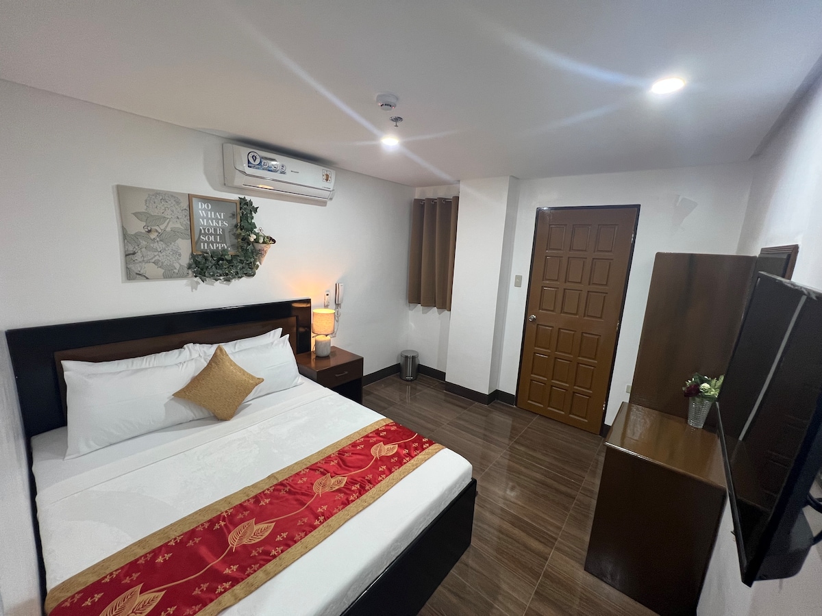 An air-conditioned room features a spacious bed dressed in crisp white linens, accented with a decorative red runner. A flat-screen television is mounted on the wall opposite the bed. There are neutral walls and wood-tone furnishings, creating a calming retreat.