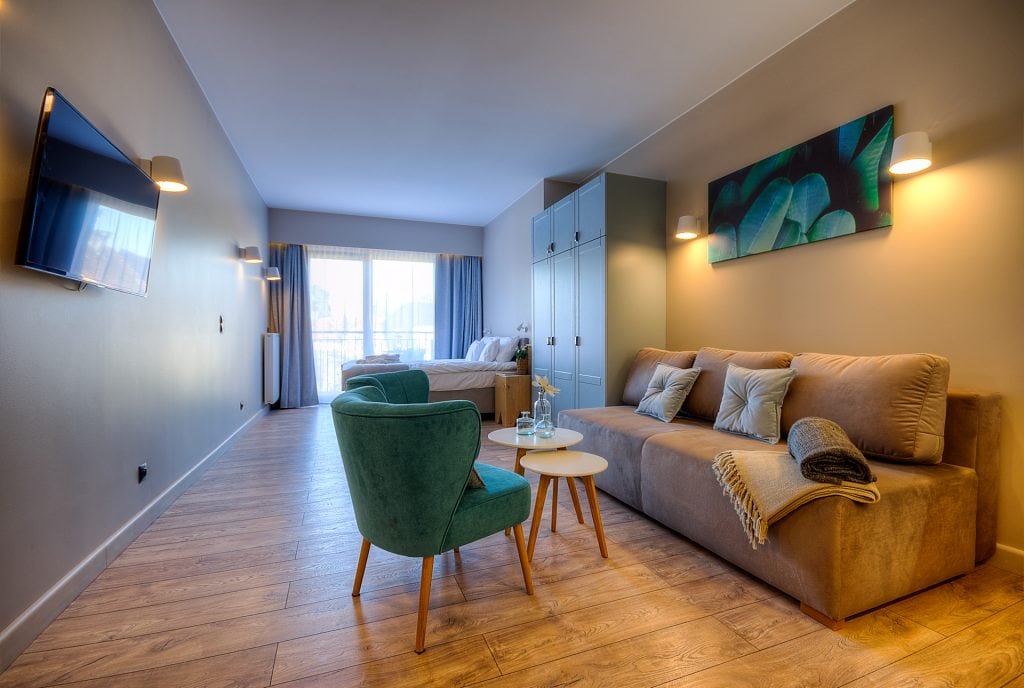 A spacious living area features a comfortable beige sofa and two green armchairs arranged around a small white table. A flat-screen TV is mounted on the wall, while large windows allow natural light to brighten the room. Soft blue and green accents are visible in the artwork above the sofa.