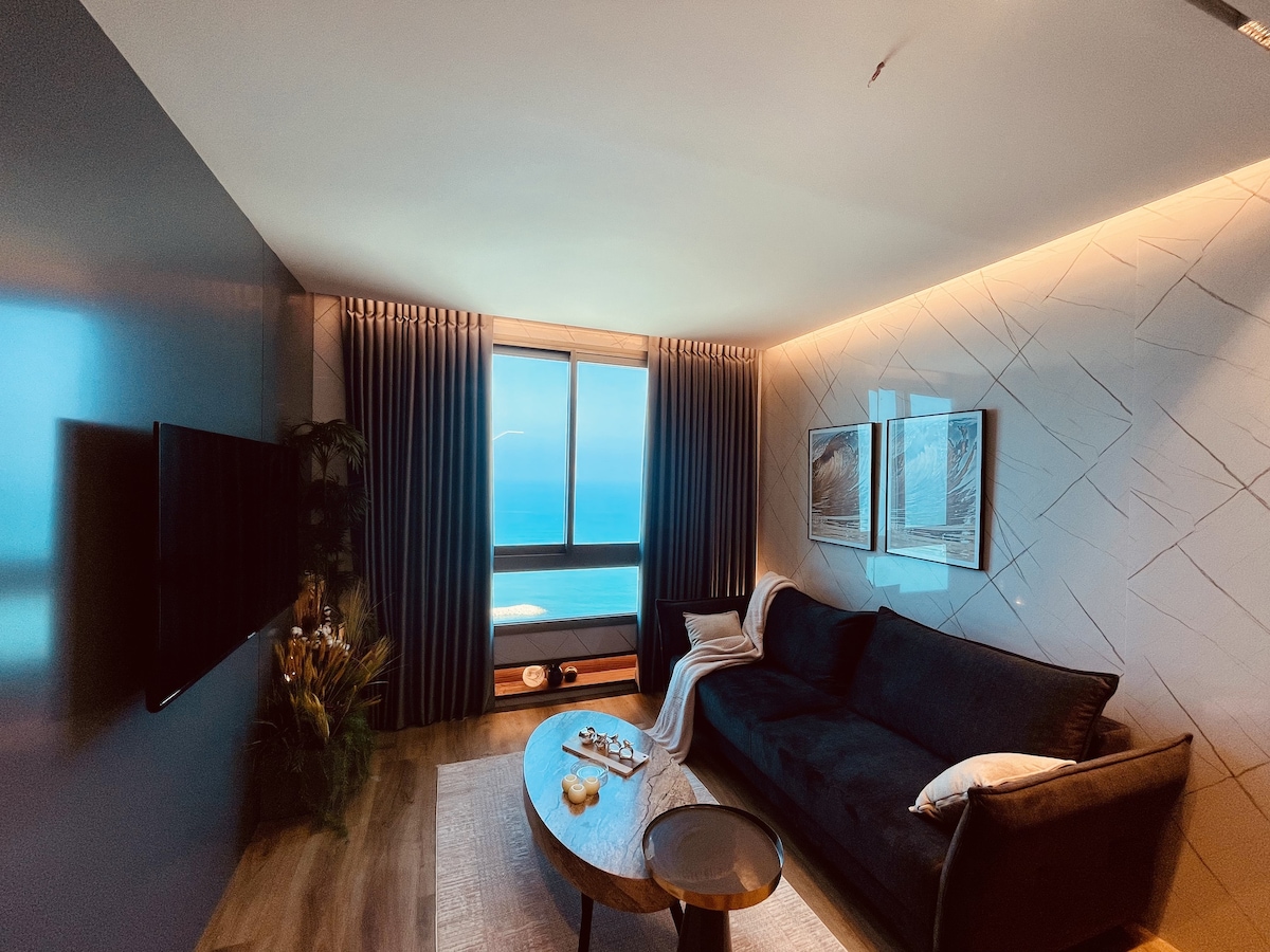 The living area features a dark sofa positioned in front of a large window, offering panoramic sea views. Light filters through long curtains, creating a calming ambiance. A round coffee table is centered, complemented by decorative items. Framed artwork adorns the wall, enhancing the space.