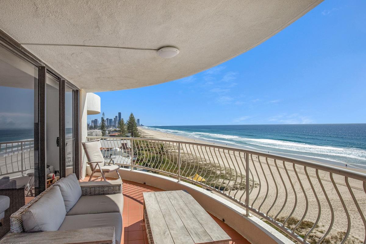 A spacious balcony offers sweeping views of the beach and ocean, with gentle waves lapping against the shore. Comfortable outdoor seating is available alongside a wooden coffee table, creating a serene space for relaxation. The city skyline can be seen in the distance.