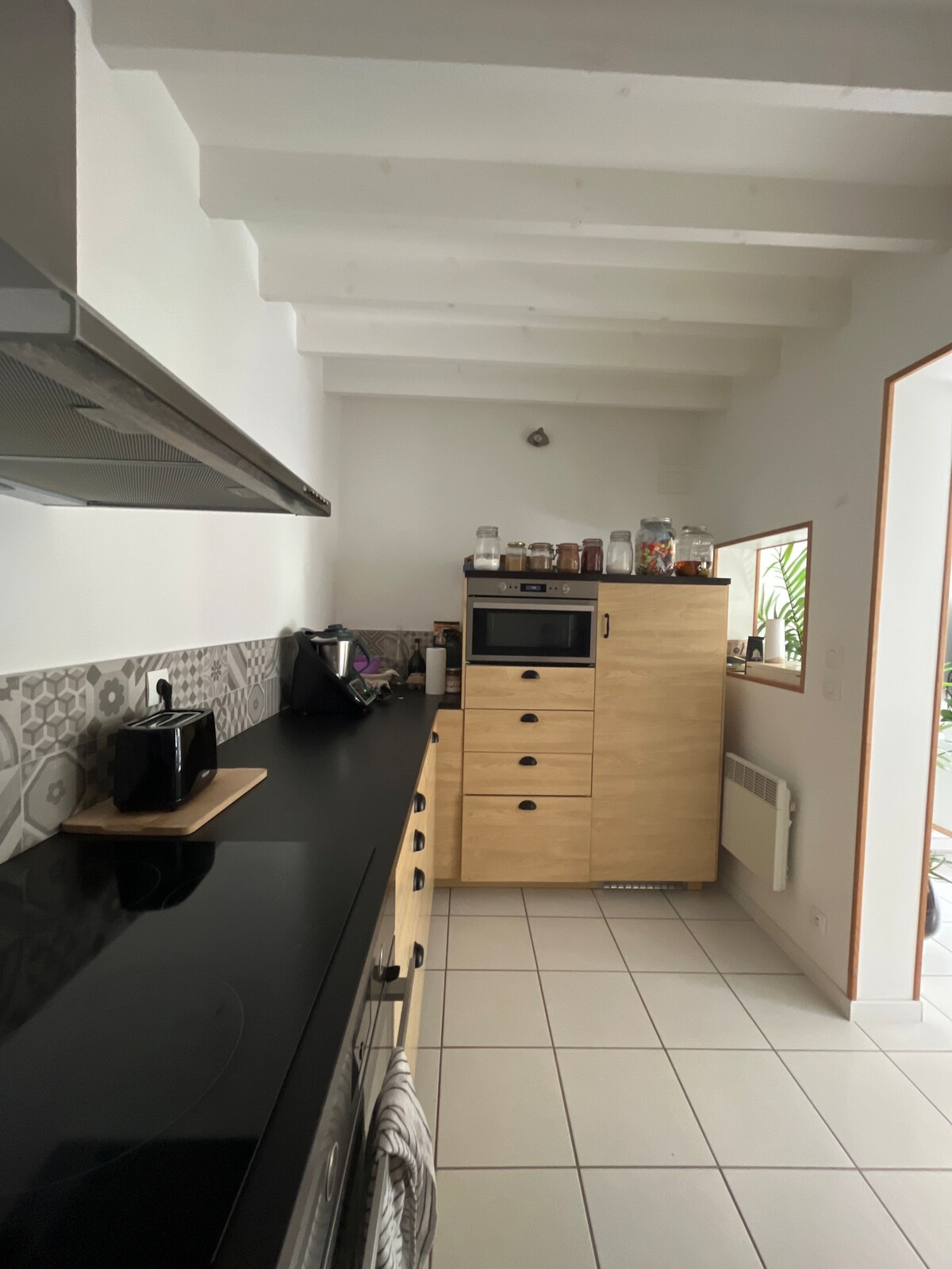 The kitchen features modern cabinetry in light wood tones, complemented by a sleek black countertop. A built-in oven and a coffee maker are visible on the countertop, while patterned tiles add visual interest to the backsplash. Natural light is provided through an adjacent opening.