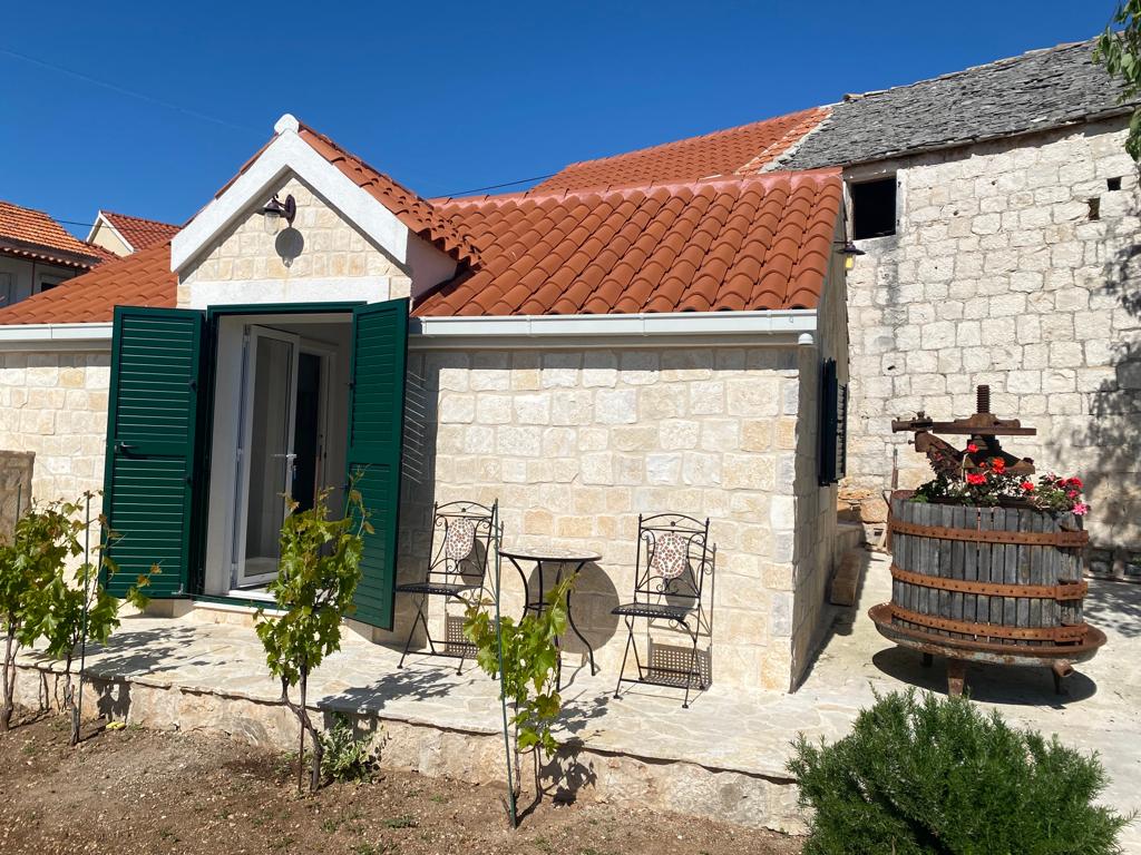A charming stone cottage is featured with green shutters, positioned against a clear blue sky. A small outdoor seating area with two wrought iron chairs and a table is visible, accompanied by a traditional wooden barrel adorned with vibrant flowers, enhancing the serene surroundings.