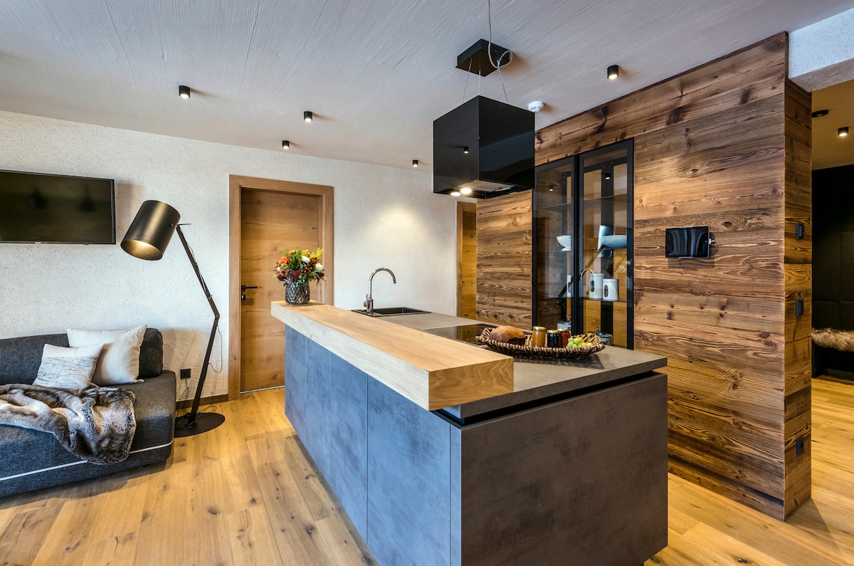 An open kitchen area features a modern island with a wooden countertop and dark cabinetry. A sleek lamp stands beside a cozy sofa, and a decorative arrangement of flowers is displayed on the kitchen island. Warm wood tones are complemented by a textured wall.