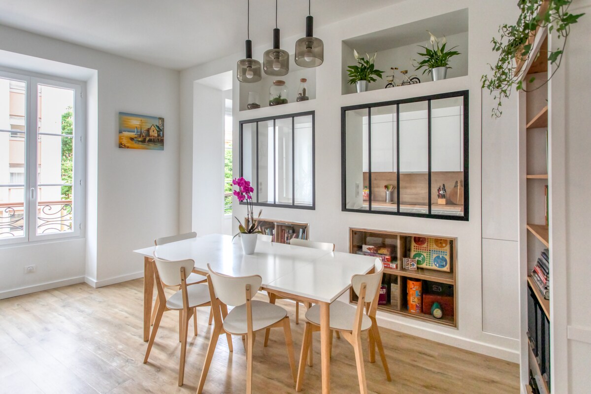A bright dining area features a spacious white table surrounded by six light wooden chairs. Above, three pendant lights provide soft illumination. Shelves are arranged with decorative plants and various objects, while large windows offer views of the outdoors.