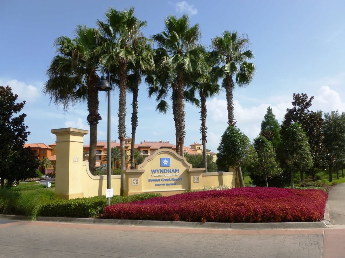 The entrance to Bonnet Creek Resort is framed by palm trees and lush shrubs, with colorful flowering plants at the forefront. A welcoming sign displays the name of the resort, complemented by a clear blue sky above.