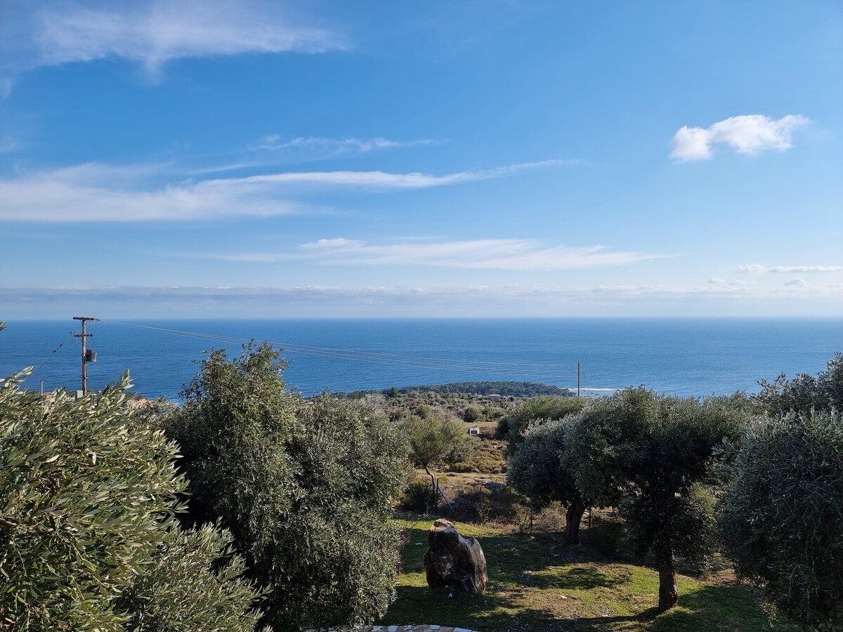 A scenic view showcases the vast expanse of the Aegean Sea, bordered by rolling hills and dotted with olive trees. The bright blue sky is punctuated with soft clouds, creating a serene atmosphere in this natural landscape.