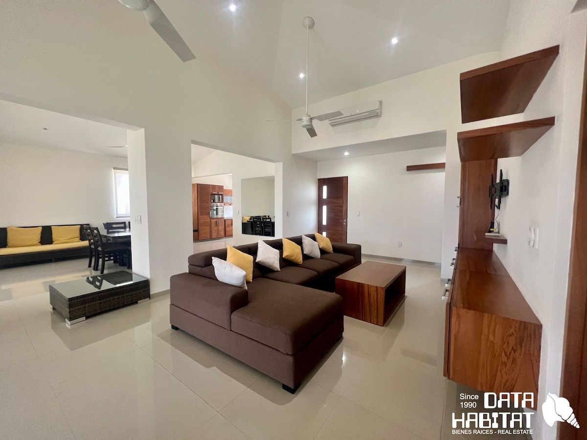 An open living area features a large sectional sofa with cushions arranged on one side. A coffee table sits in front of the sofa, and a cabinet is visible against the wall. Soft natural lighting enters through windows, enhancing the spacious ambiance.