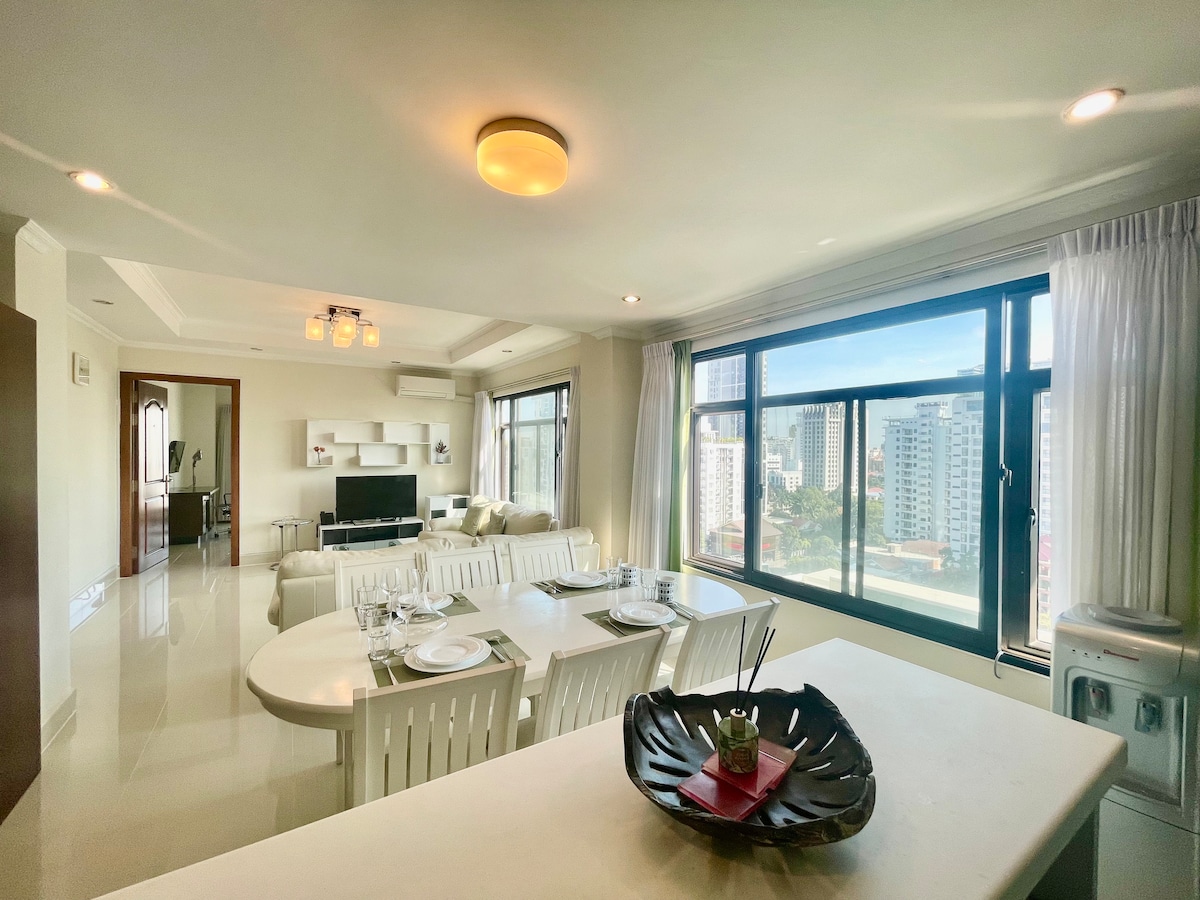A bright living space features large windows that provide expansive views of the city skyline. A modern dining area with a white table and chairs is complemented by a sleek kitchen counter. Comfortable seating is arranged for relaxation, and natural light enhances the ambiance.
