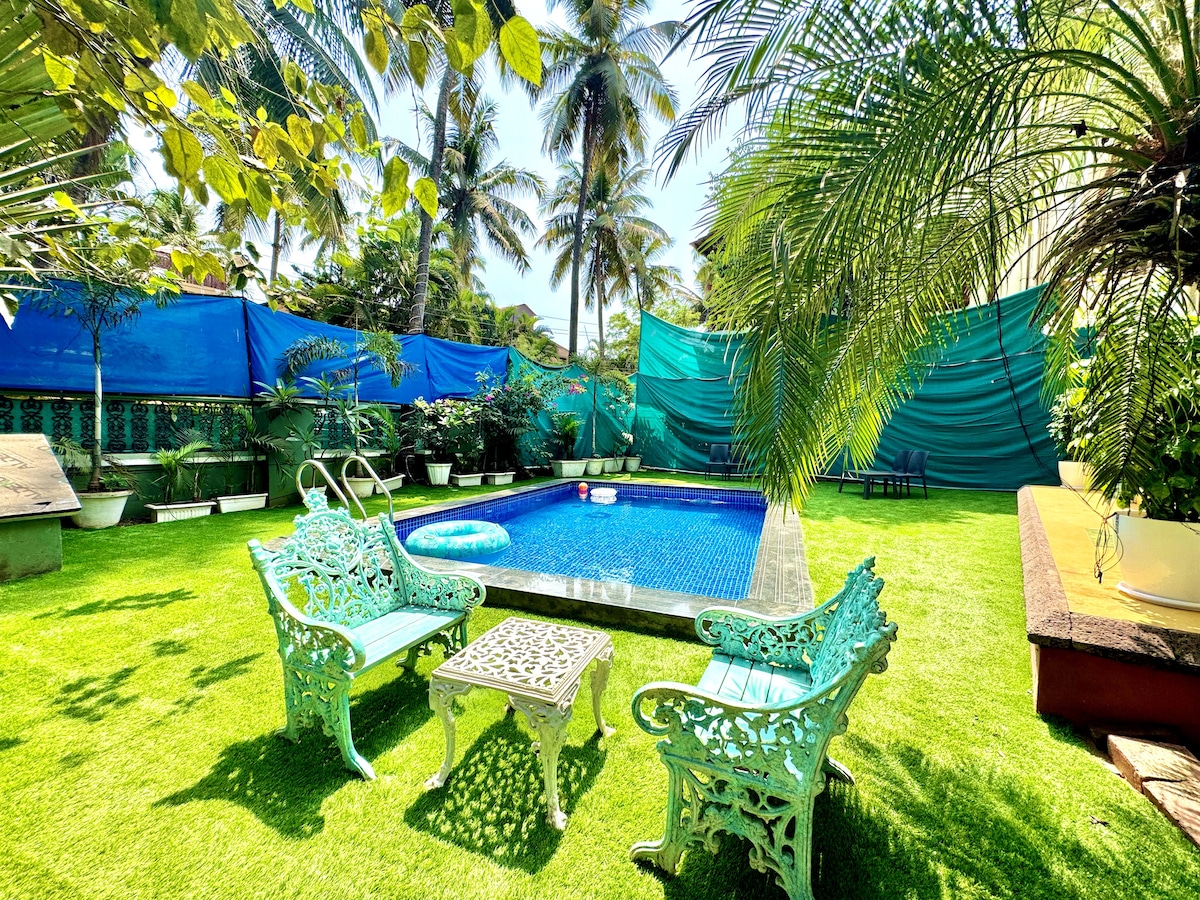 An outdoor space featuring a private swimming pool set against vibrant greenery. Lounge chairs with intricate designs surround the pool, complemented by a small table. Tall palm trees and lush plants create a tranquil atmosphere while blue tarps provide shade in the background.