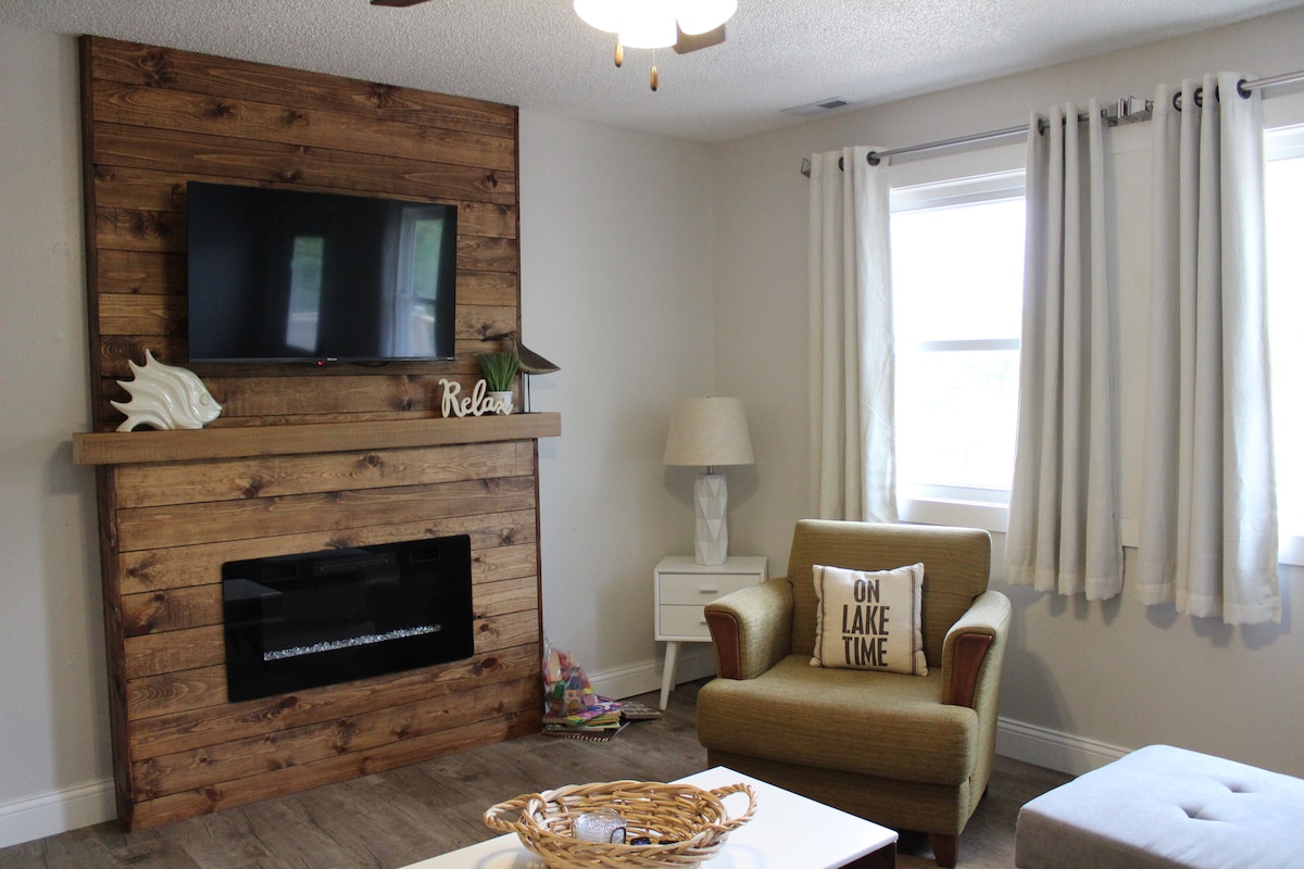 A cozy living area is highlighted by a wooden accent wall, featuring a mounted flat-screen TV above a gas fireplace. An armchair with a decorative pillow sits beside a small side table and lamp, while light curtains frame the window, allowing natural light to enter.