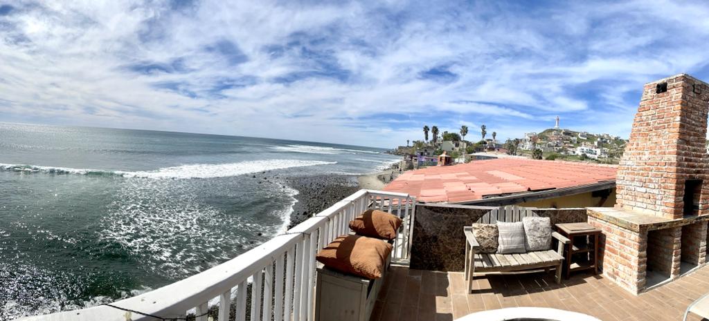 A panoramic view of the ocean is displayed from a spacious outdoor terrace. Comfortable seating is arranged nearby, overlooking gentle waves and a rocky shoreline. A stone barbecue grill is positioned beside the seating area, enhancing the outdoor living experience.