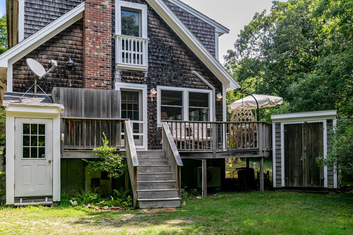 The rear exterior of a two-story cottage is depicted, featuring a wooden deck and stairs leading to the ground. Two outdoor structures are visible, one resembling a small shed and another with a door. Lush greenery surrounds the area, contributing to a natural setting.