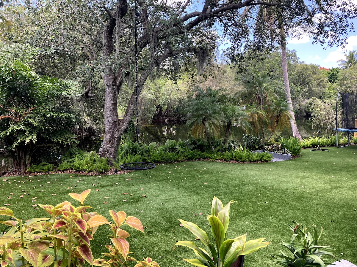A lush backyard features well-maintained greenery and vibrant plants. A manicured lawn of artificial grass provides a serene area. Tall trees and shrubs surround a calm lake, enhancing the natural landscape.