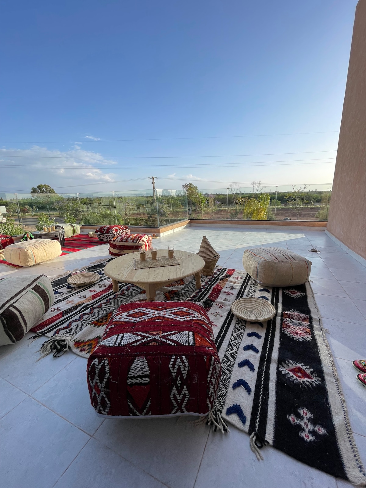 A spacious terrace features traditional Moroccan decor, including colorful poufs and crafted wooden furniture. A large round table stands at the center, surrounded by patterned rugs. Expansive glass panels provide an unobstructed view of the greenery and distant horizon.