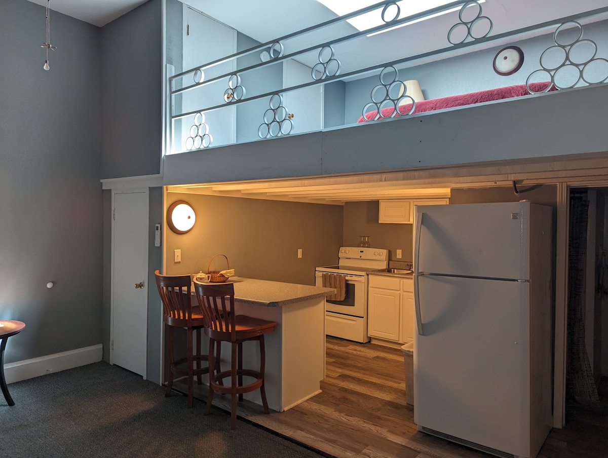 The open living area features a kitchen with modern appliances, including a refrigerator and stove. A breakfast bar with two stools provides seating. Above, a lofted bedroom area is visible, complemented by a thoughtfully designed railing and a skylight that allows natural light to enter.