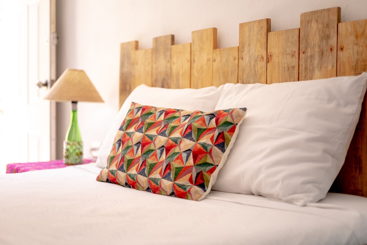 A well-made bed is presented with crisp white linens and a decorative pillow featuring a colorful geometric pattern. A wooden headboard adds a rustic touch, while a bedside lamp and a vibrant tablecloth introduce additional color and texture to the space.