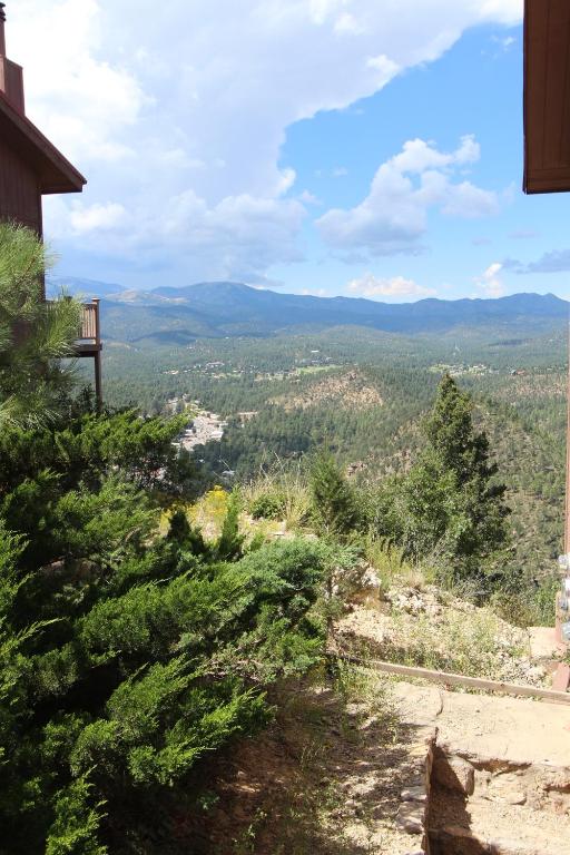A scenic view of mountainous terrain stretches across the horizon, featuring lush greenery and rolling hills. Clear blue skies are dotted with clouds, while the distant peaks stand majestically. The foreground includes dense shrubs, adding a natural framing to the expansive landscape.