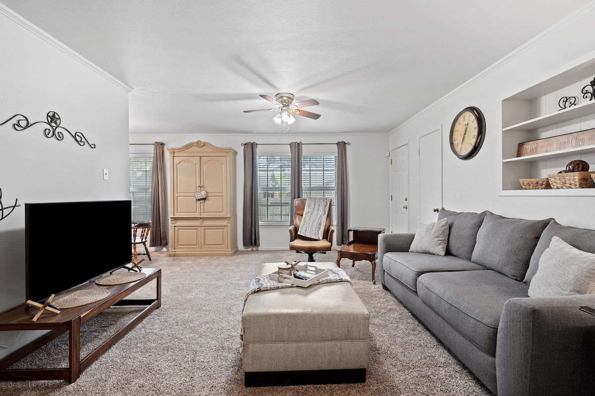 The living area features a large gray sofa positioned near a plush ottoman, providing ample seating. A flat-screen television is placed on a wooden stand, while natural light enters through multiple windows, enhancing the bright and open feel. A tasteful wall clock and decorative shelving add to the decor.